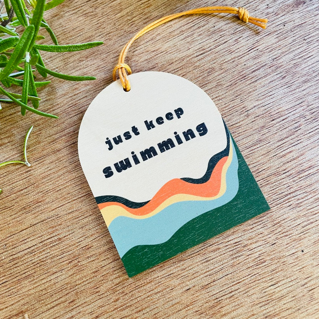 Wooden hanging decoration with the wording 'Just Keep Swimming' with colourful waves