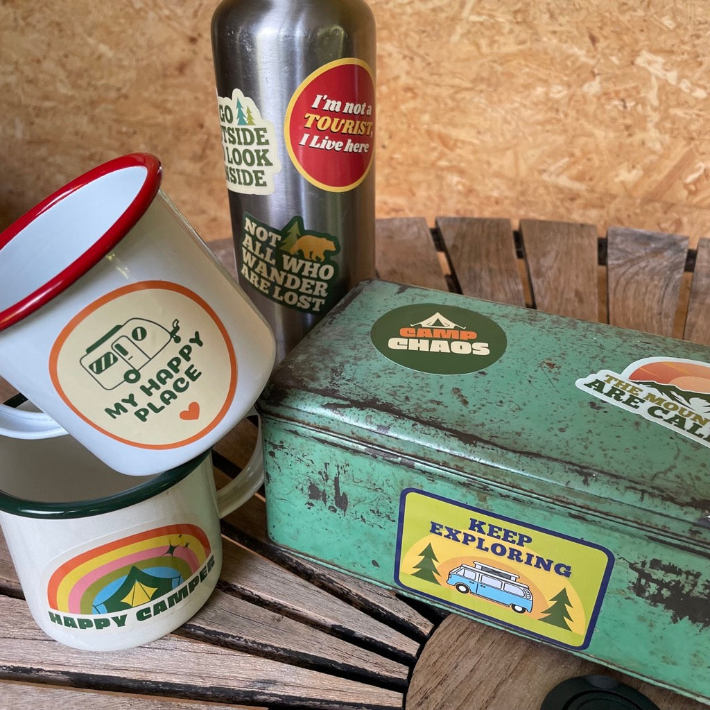 Various adventure stickers displayed on a water bottle, mugs, and tin.