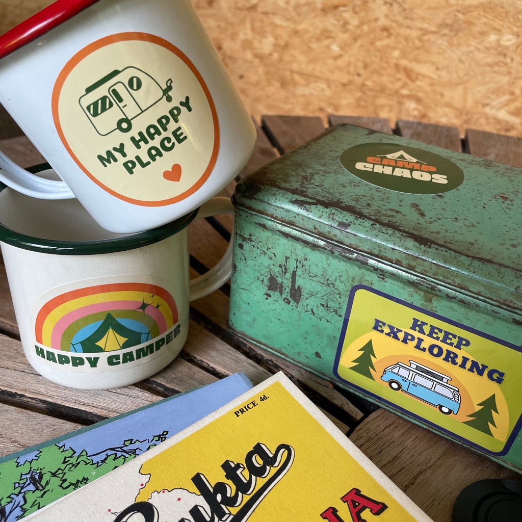 Various adventure stickers displayed on a water bottle, camping mugs and tin.