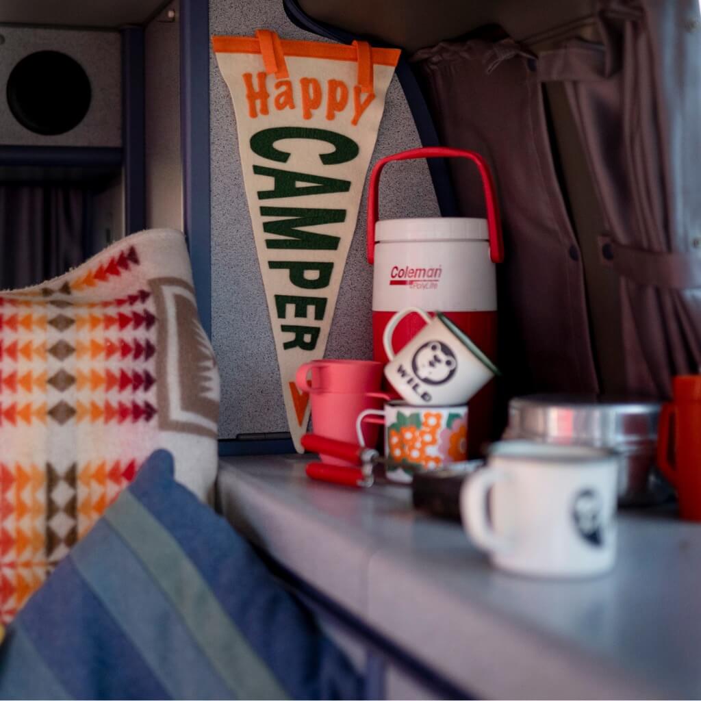 Happy Camper flag hanging in a campervan with enamel mugs and retro flask