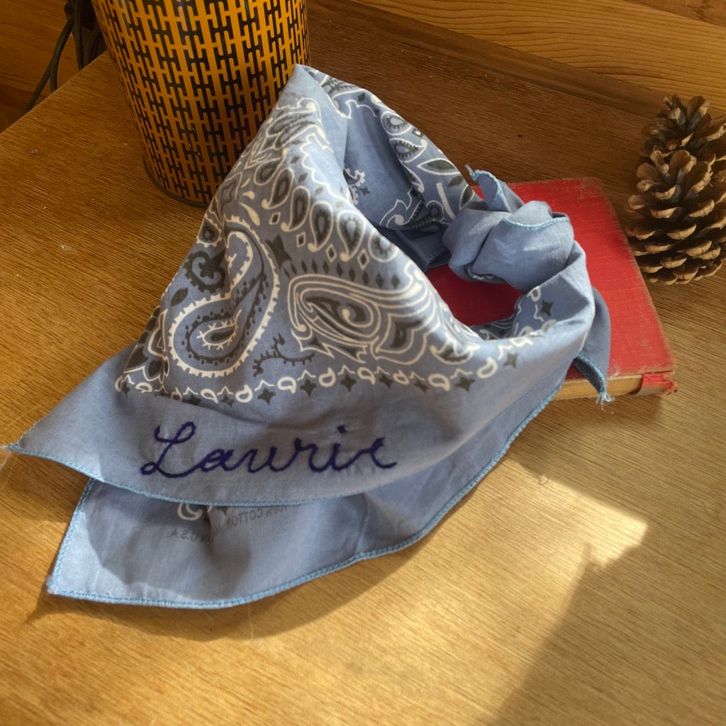 A custom chain stitched bandana in bluesitting on a desk with pine cones and a camping mug.