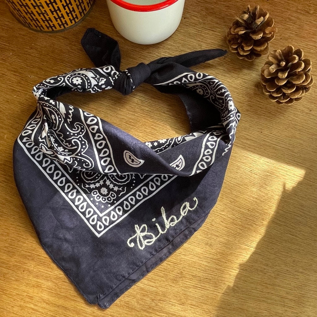 A custom chain stitched bandana in navy sitting on a desk with pine cones and a camping mug.