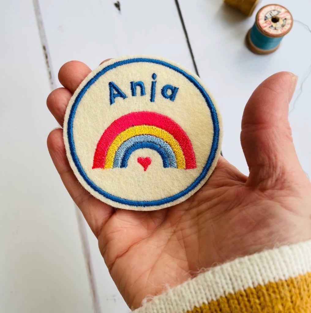 Rainbow  embroidered name patch in cream and blue being held in a hnad for scale