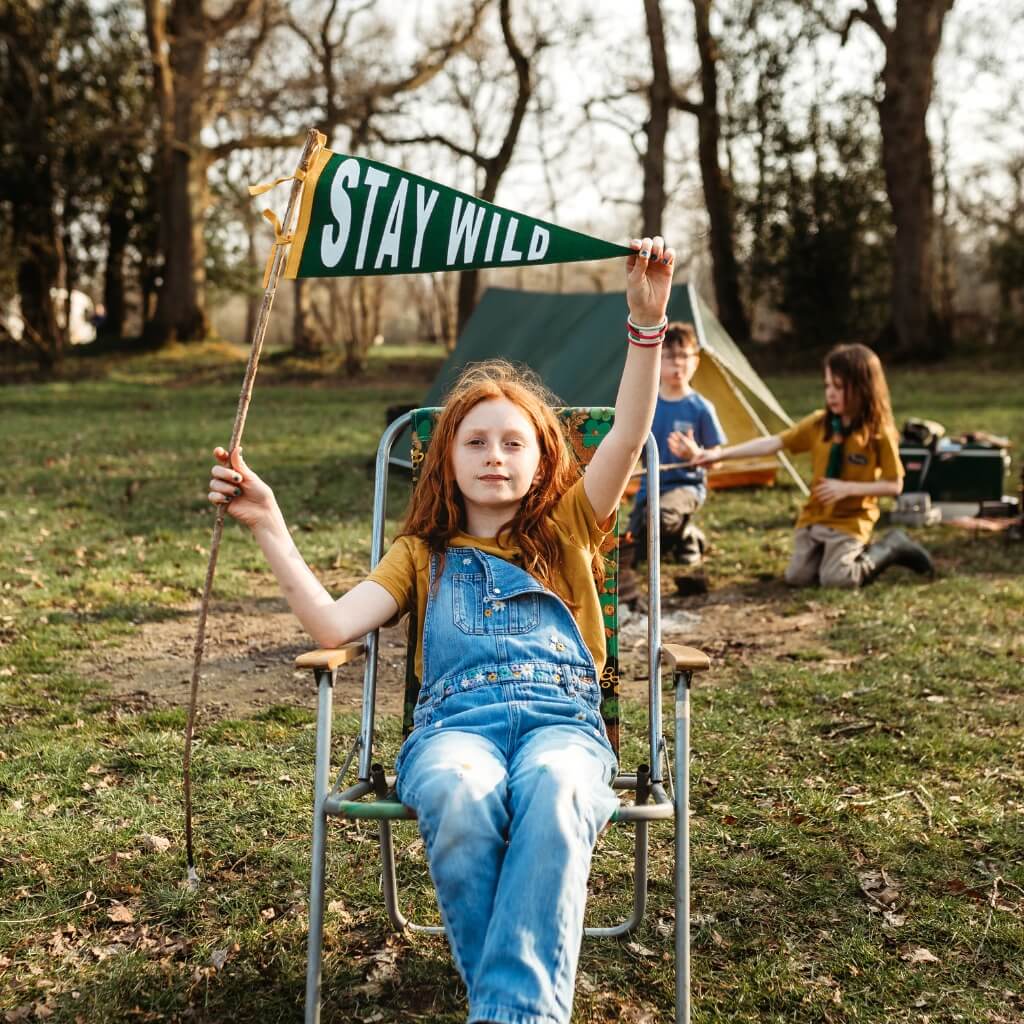 A girl with red hair sitting in a deck chair holding up a stay wild pennant flag